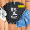 May include: Black t-shirt with white text that reads "Sorry I'm Late Lucy Was Sitting On Me" with a white silhouette of a dog sitting on the text. The shirt is laid out on a wooden surface with a pair of white sneakers and blue jeans.