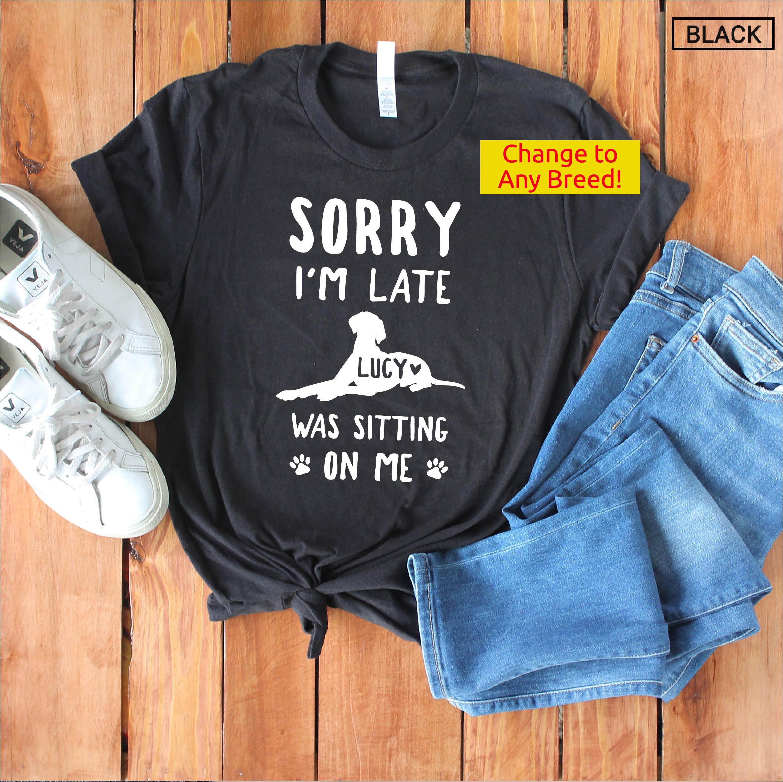 May include: Black t-shirt with white text that reads "Sorry I'm Late Lucy Was Sitting On Me" with a white silhouette of a dog sitting on the text. The shirt is laid out on a wooden surface with a pair of white sneakers and blue jeans.